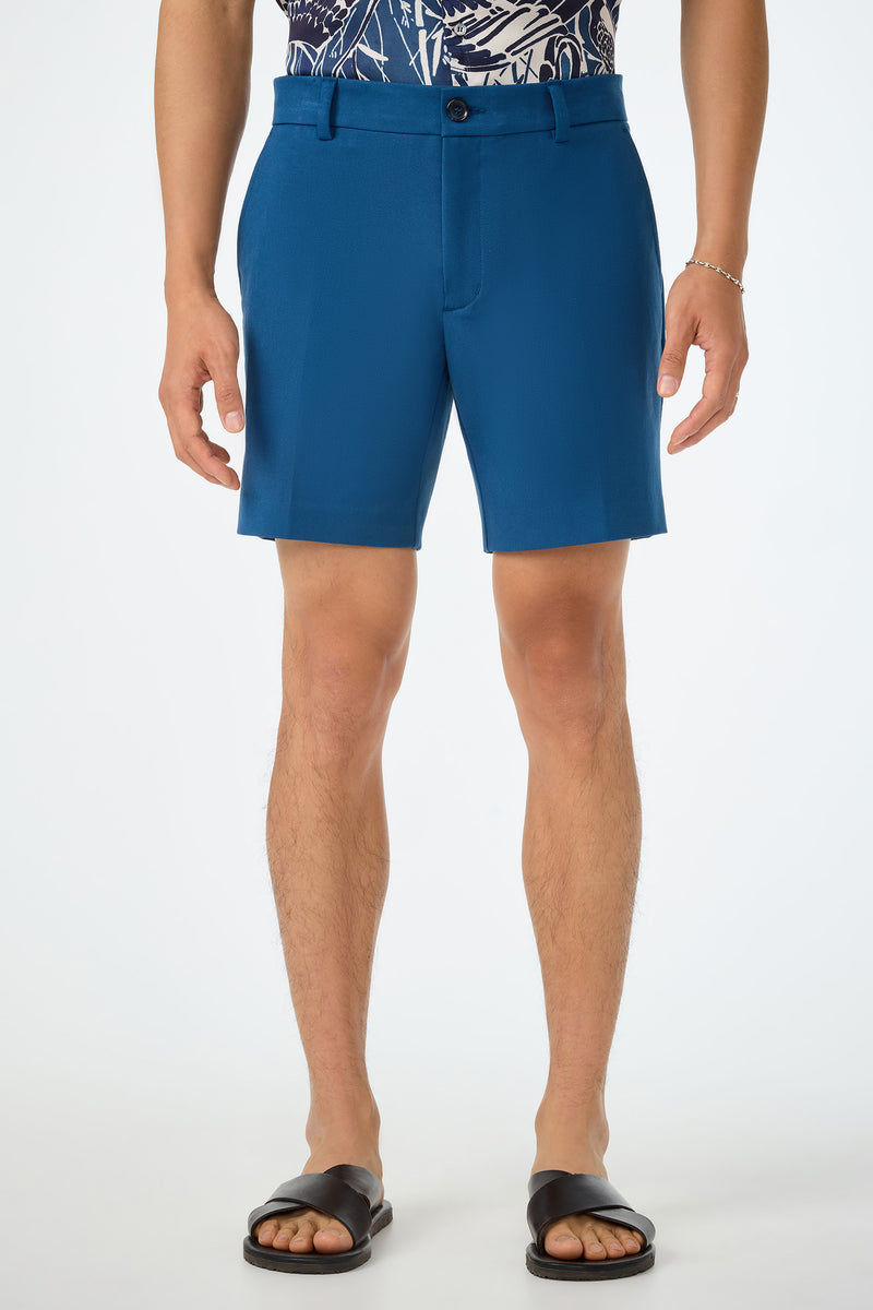 A person wearing the Travis Short in Sumi Blue, paired with a navy patterned shirt, black slide sandals, and a silver bracelet, stands against a plain white background.