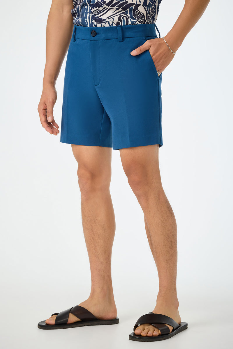 A person wears the Travis Short - Sumi Blue, paired with a patterned navy shirt and black cross-strap sandals, standing against a plain white background. Only their lower half is visible.