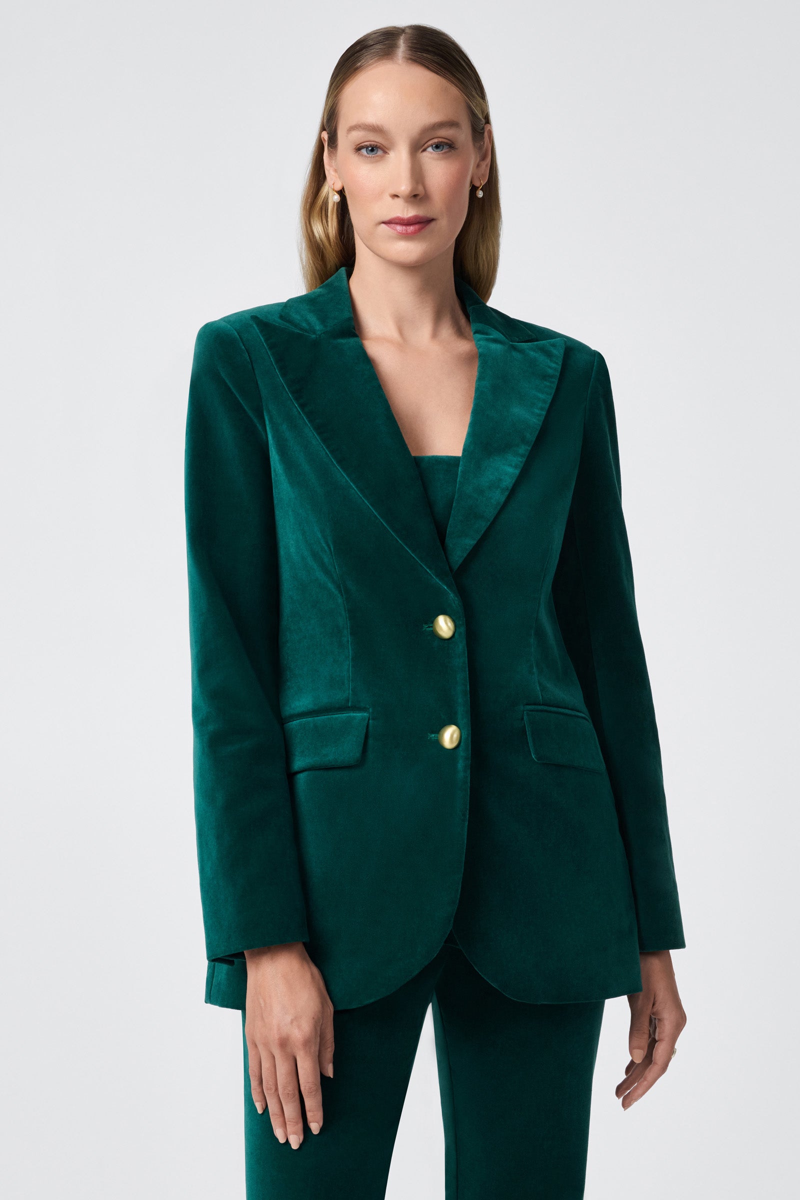 A woman with light brown hair models the Broadway Jacket, a deep green velvet suit with gold buttons, against a plain light gray background.