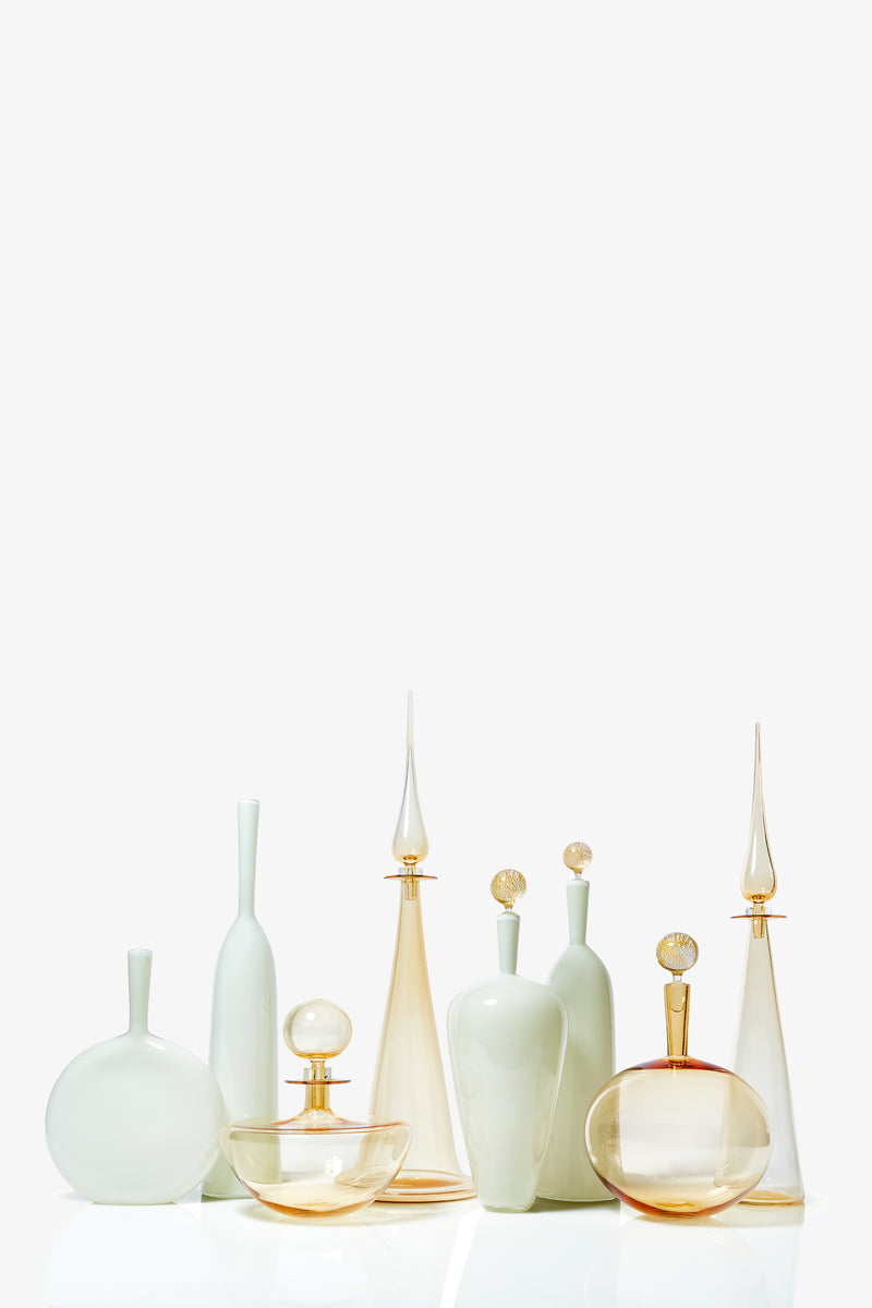 A set of Carmella Barware Low Round glass bottles and vases in soft white and pale amber, featuring different heights and shapes, arranged together against a plain white background.