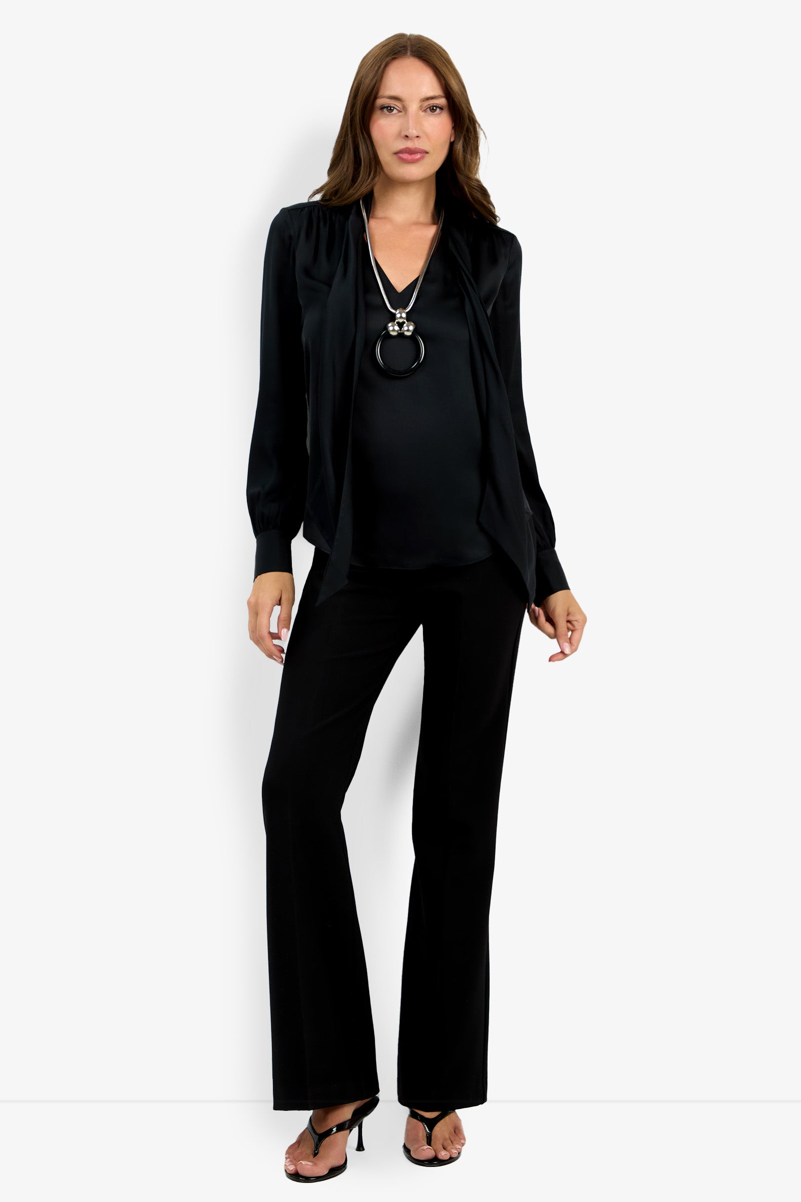 A woman with straight brown hair and a neutral expression stands against a light background wearing the Deming Top, black wide-leg pants, heeled sandals, and a long silver necklace.