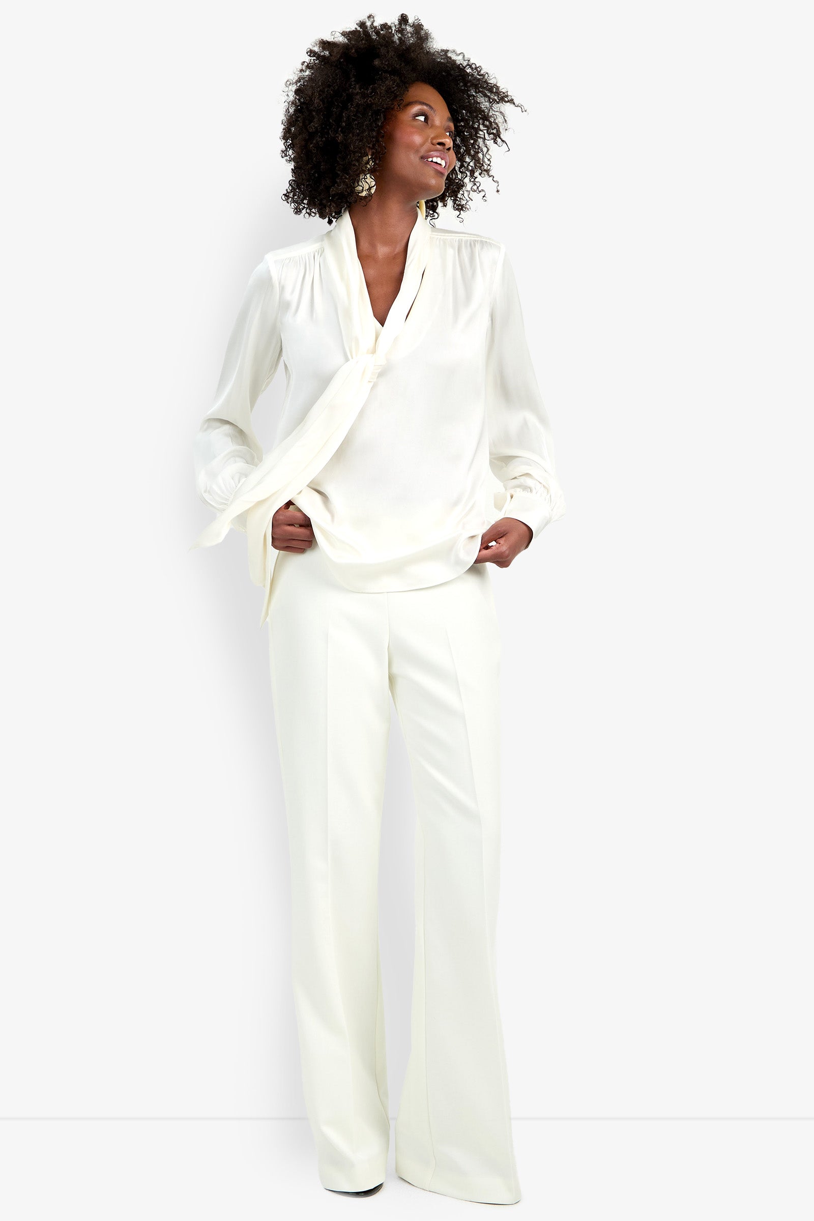 A woman smiles while wearing the Deming Top—an elegant ivory blouse with a tie-neck detail—and matching wide-leg trousers, posing against a plain white background.