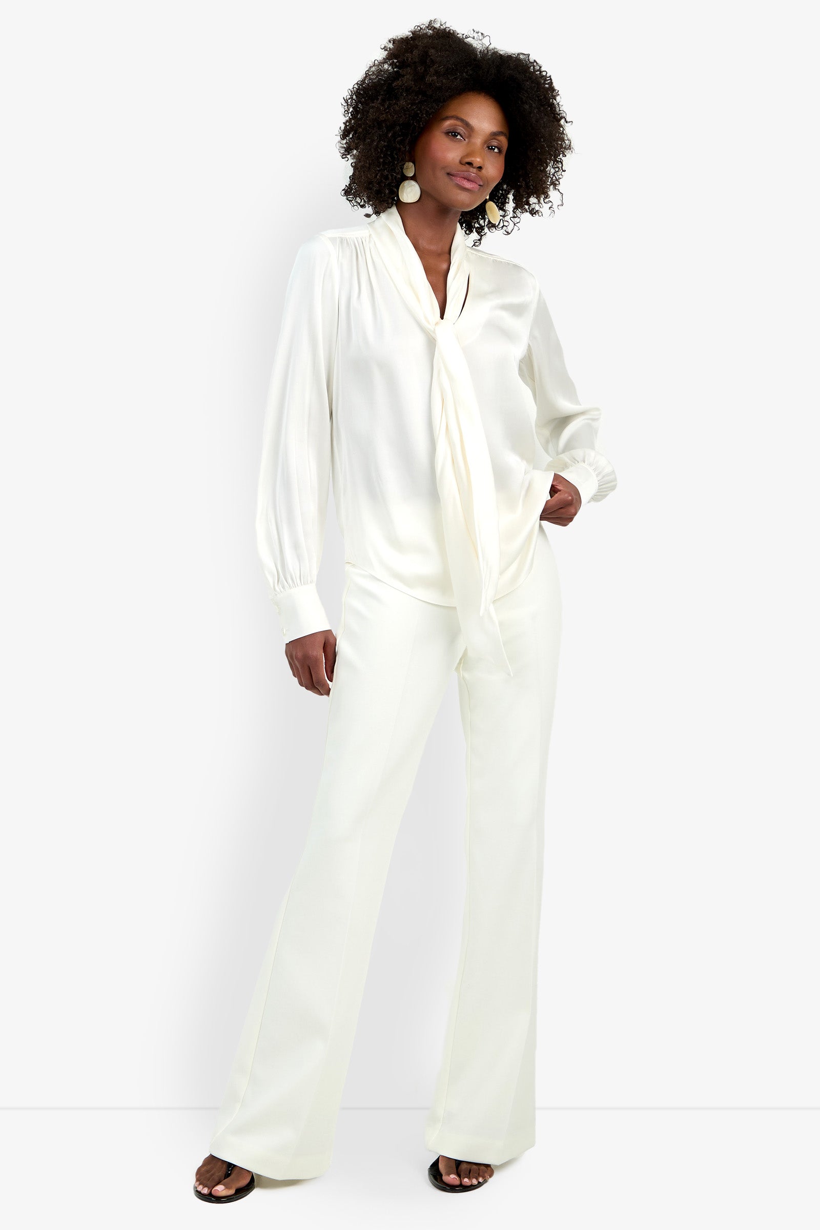 A woman stands confidently in the Deming Top, a silky white blouse with a tie-neck detail, paired with matching wide-leg trousers. She has natural curly hair, large round earrings, and black open-toe shoes against a plain background.