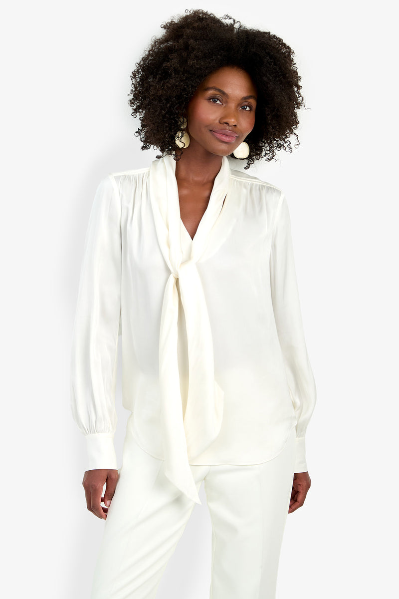 A woman with curly hair wears the Deming Top, a white blouse with a long necktie, paired with white pants. She stands against a plain white background, smiling softly at the camera.