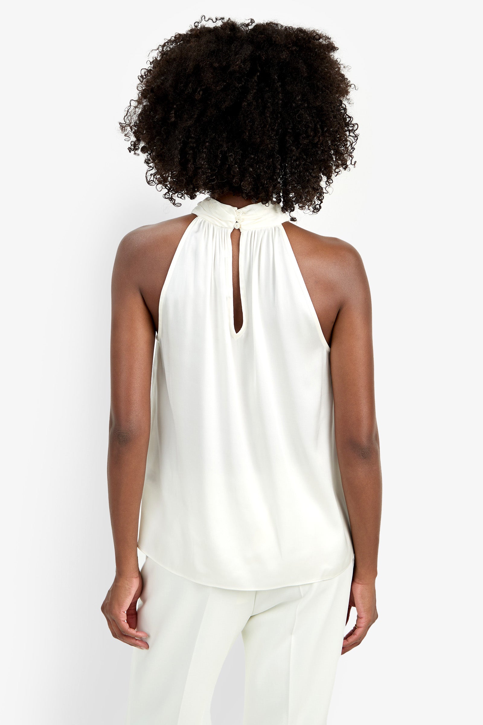 A woman with curly hair is shown from the back, wearing the sleeveless white Zana Top with a keyhole neckline and white pants against a plain white background.