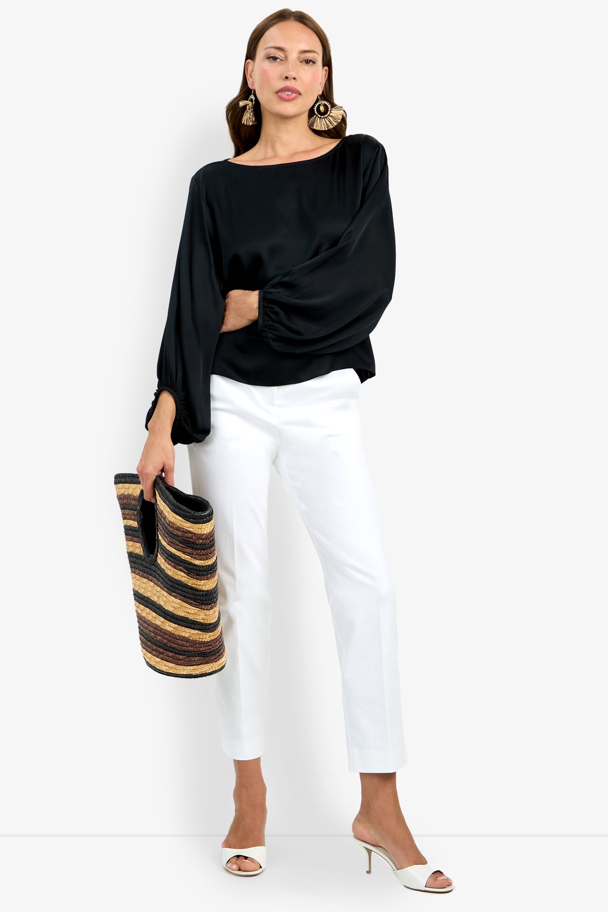 A woman wearing the Ursa Top, black long-sleeve blouse, white pants, and white heeled sandals holds a striped woven tote bag. She accessorizes with large statement earrings and stands against a plain background.