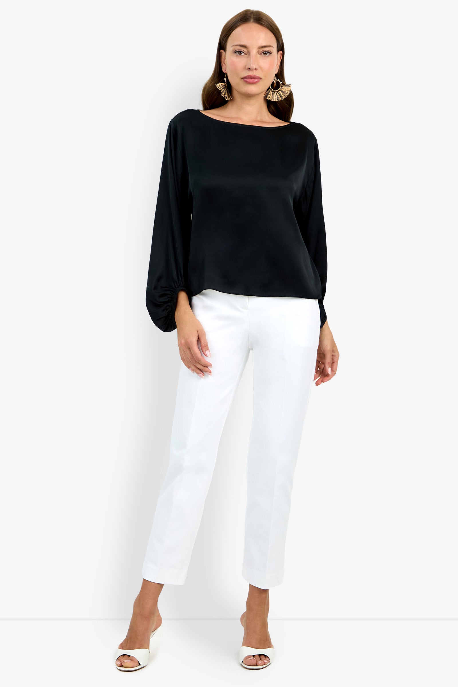 A woman with straight hair and a neutral expression stands against a white background wearing the Ursa Top, a long-sleeved black blouse, paired with white pants, white open-toe heels, and statement tassel earrings.