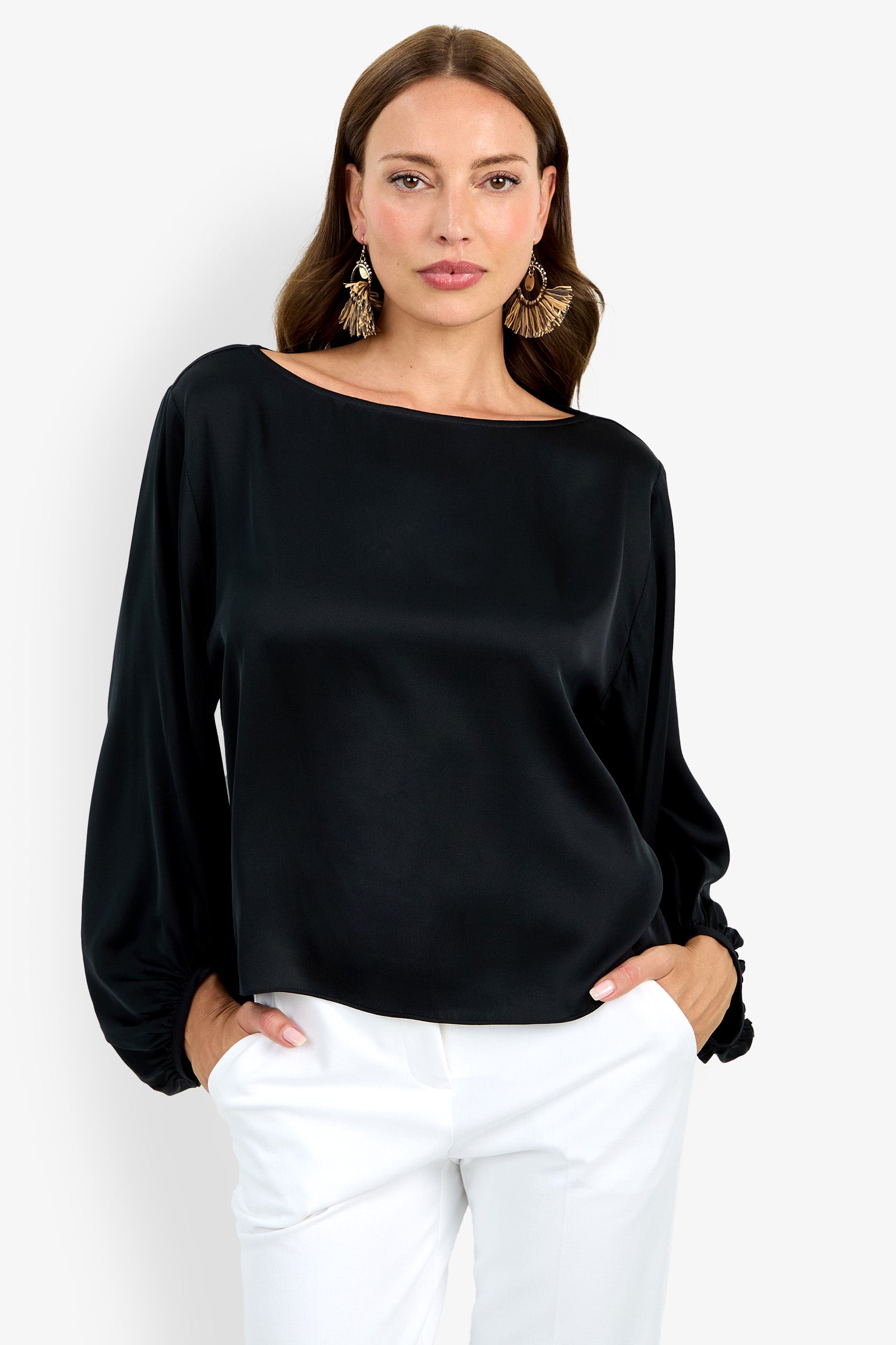 Woman with long brown hair wears statement earrings, white pants, and the Ursa Top—a loose-fitting black satin blouse with long sleeves—standing hands in pockets against a plain white background.
