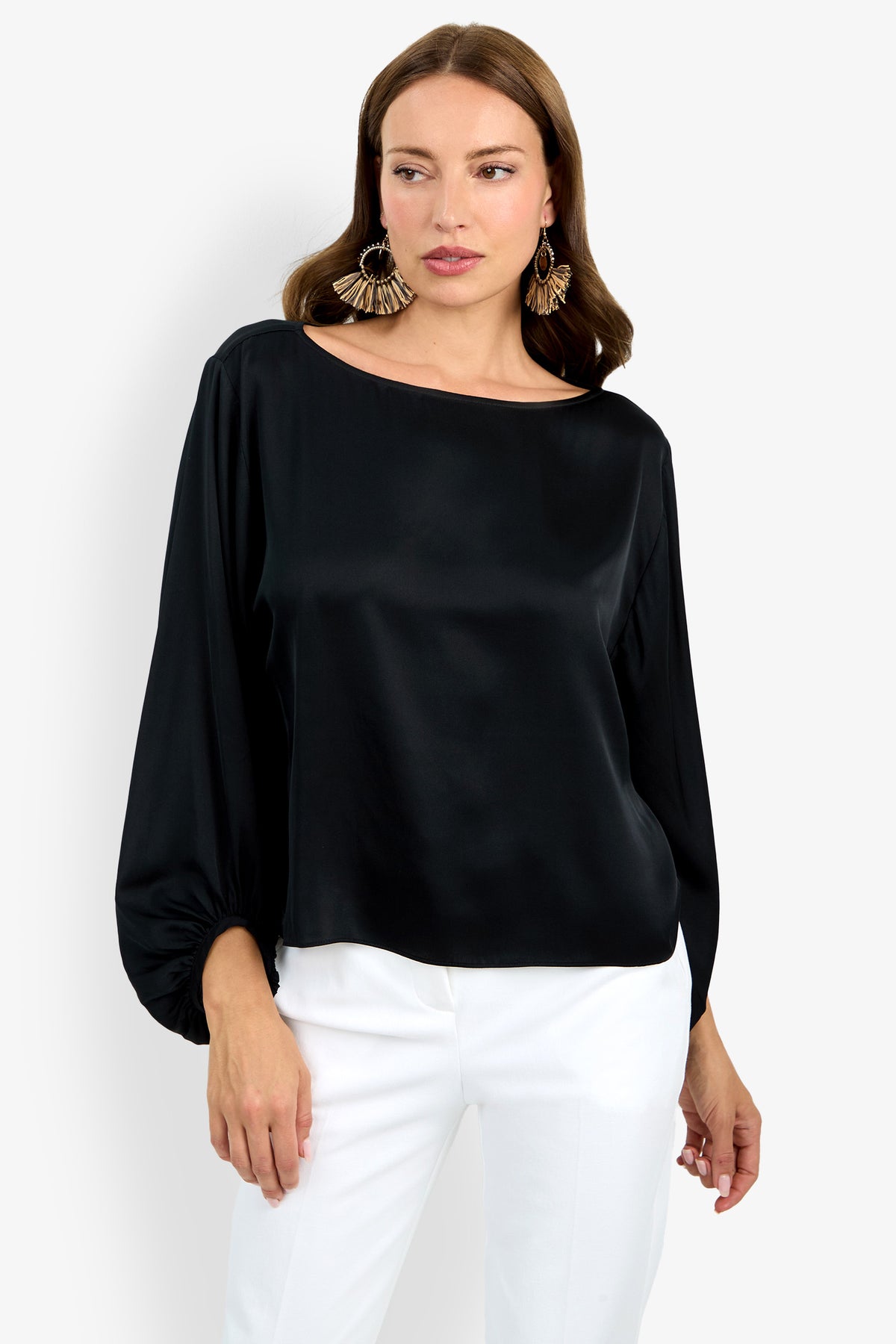 A woman with long brown hair wears the Ursa Top, a loose, long-sleeved black satin blouse, paired with white pants and large statement earrings. She stands against a plain white background.