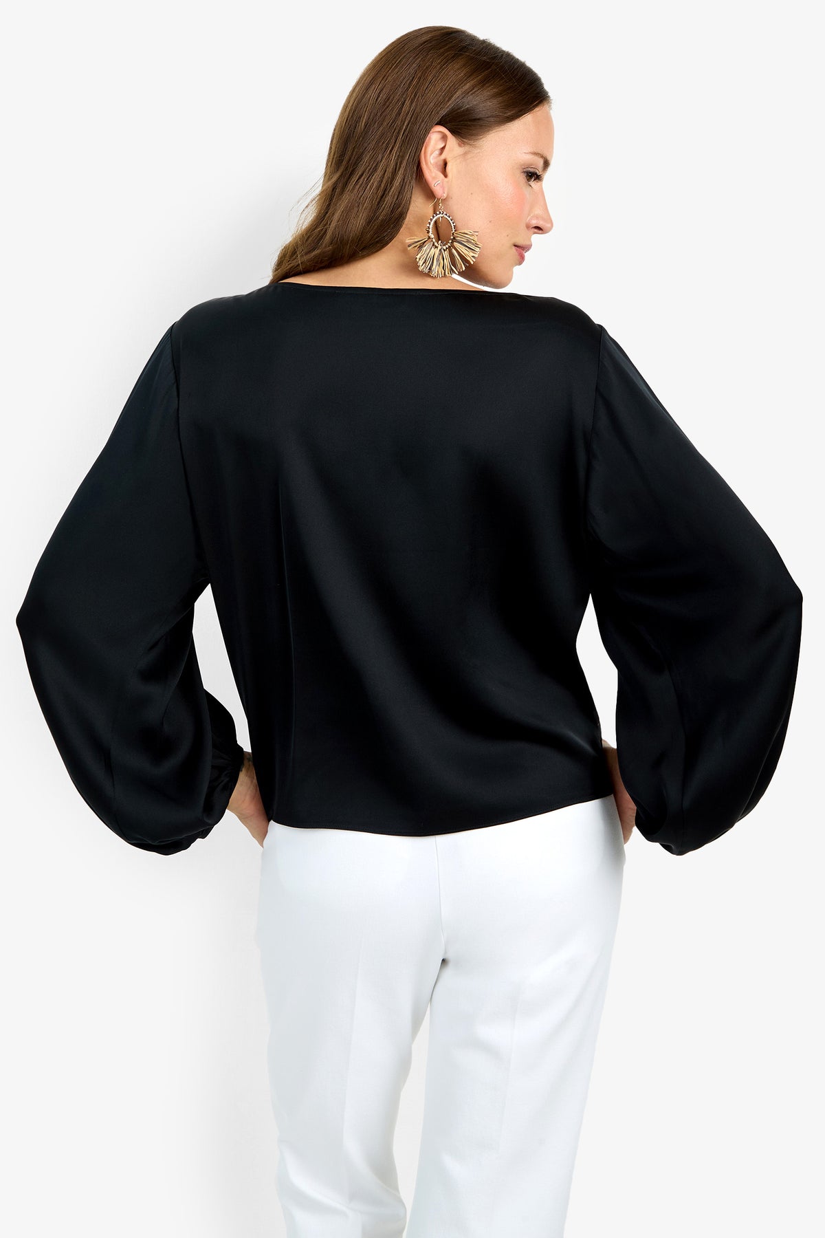 A woman with long brown hair, wearing large statement earrings and the loose-fitting black Ursa Top, stands in white pants facing away, her head turned to the side, against a plain white background.