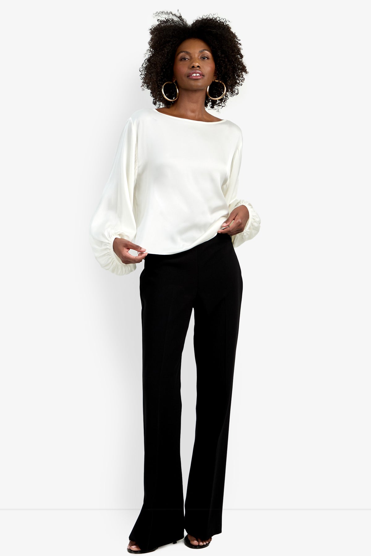 A woman stands confidently in the Ursa Top, a white long-sleeve blouse with billowy sleeves, paired with black tailored trousers, hoop earrings, and black open-toe heels against a plain white background.