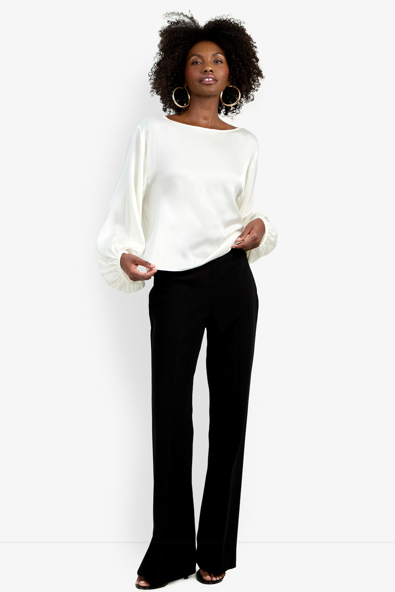 A woman stands confidently in the Ursa Top, a white long-sleeve blouse with billowy sleeves, paired with black tailored trousers, hoop earrings, and black open-toe heels against a plain white background.