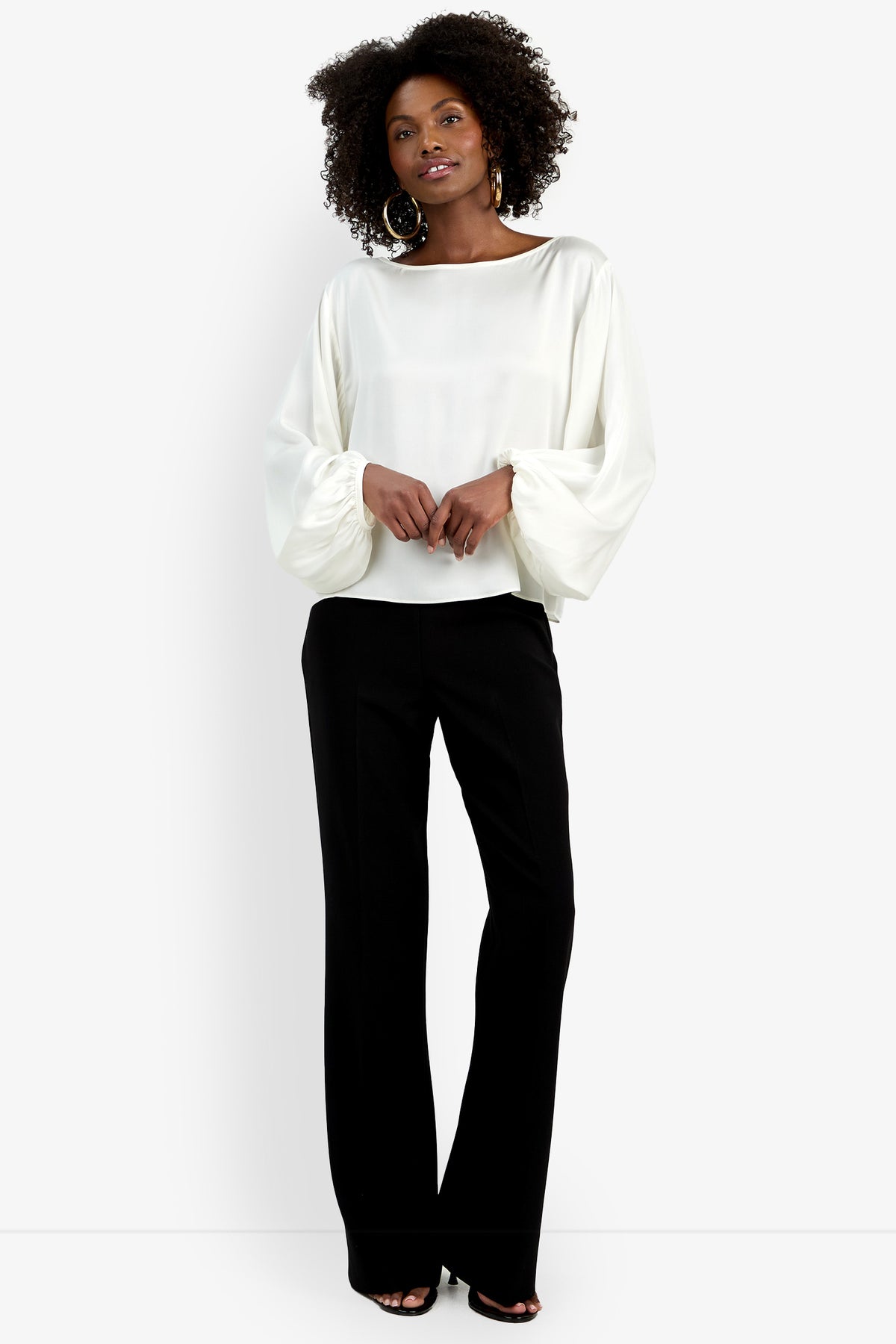 A woman with curly hair and large hoop earrings poses against a plain white background wearing the Ursa Top—a white, long-sleeve blouse with puffed sleeves—paired with black wide-leg pants.