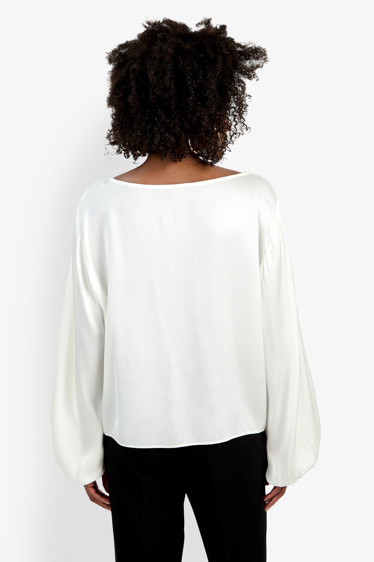 A person with curly hair is shown from the back wearing the Ursa Top, a loose-fitting, long-sleeved white blouse, paired with black pants against a plain white background.