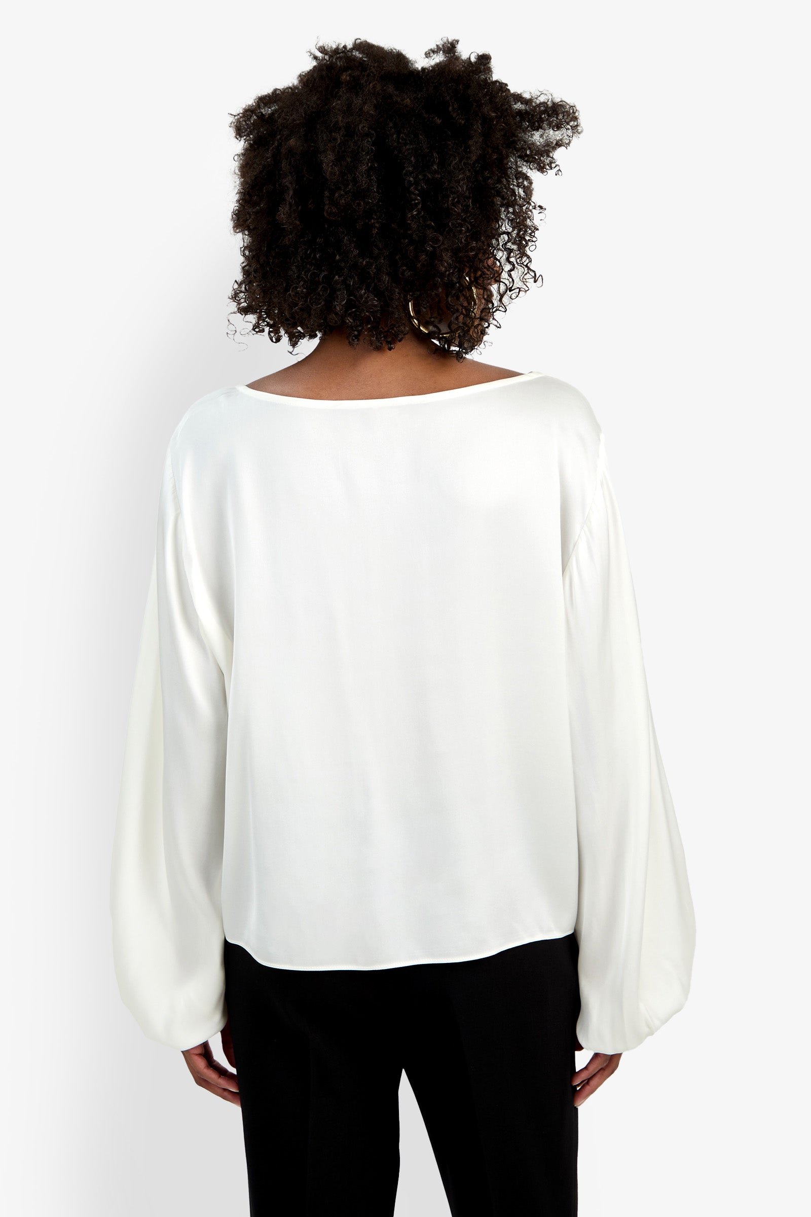A person with curly hair is shown from the back wearing the Ursa Top, a loose-fitting, long-sleeved white blouse, paired with black pants against a plain white background.
