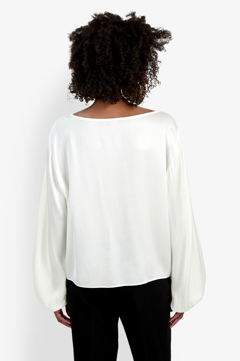 A person with curly hair is shown from the back wearing the Ursa Top, a loose-fitting, long-sleeved white blouse, paired with black pants against a plain white background.