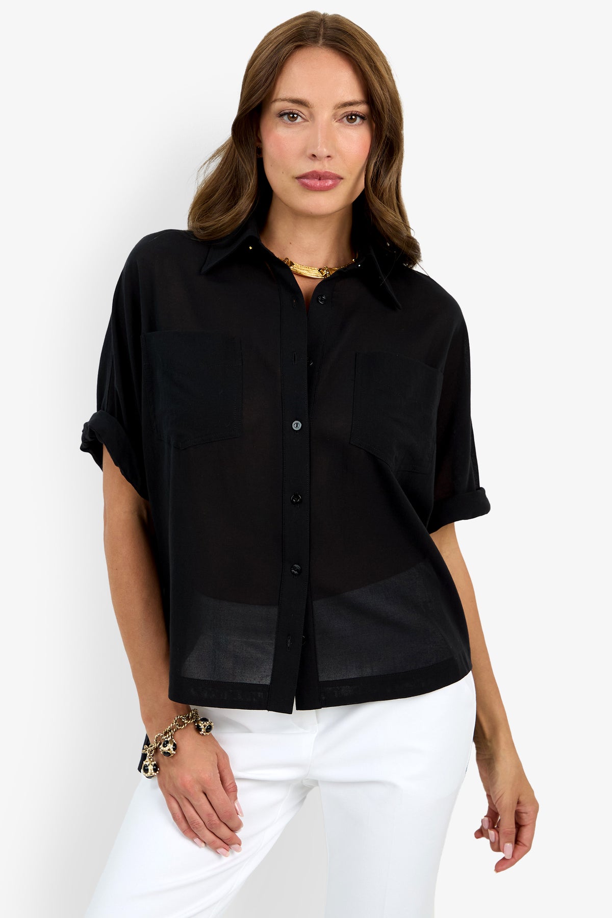 A woman with light brown hair poses confidently in the Hawaii Top—a loose-fitting, short-sleeve black button-up blouse with two front pockets—paired with white pants against a plain white background.