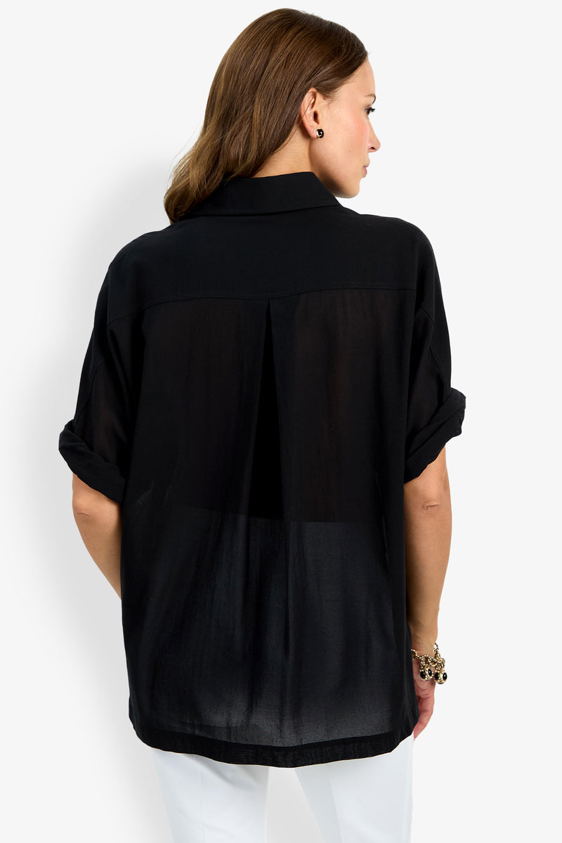 A woman with light brown hair is seen from behind, wearing the loose-fitting, black sheer Hawaii Top with rolled-up sleeves and white pants, standing against a plain white background.