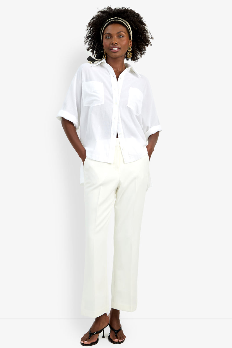 A woman stands confidently in the Hawaii Top—a loose white button-up shirt with rolled sleeves—paired with white wide-leg trousers, black heeled sandals, gold statement earrings, and a striped headband in her curly hair.
