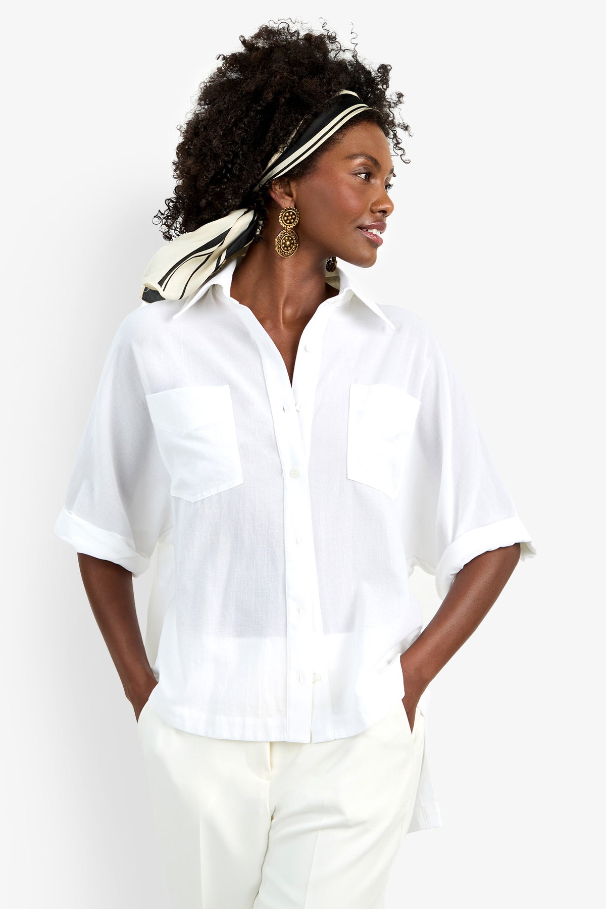 A woman wears the Hawaii Top—a loose white short-sleeve button-up with front pockets—paired with white pants. She has curly hair styled with a black-and-white scarf and accessorizes with large round earrings.