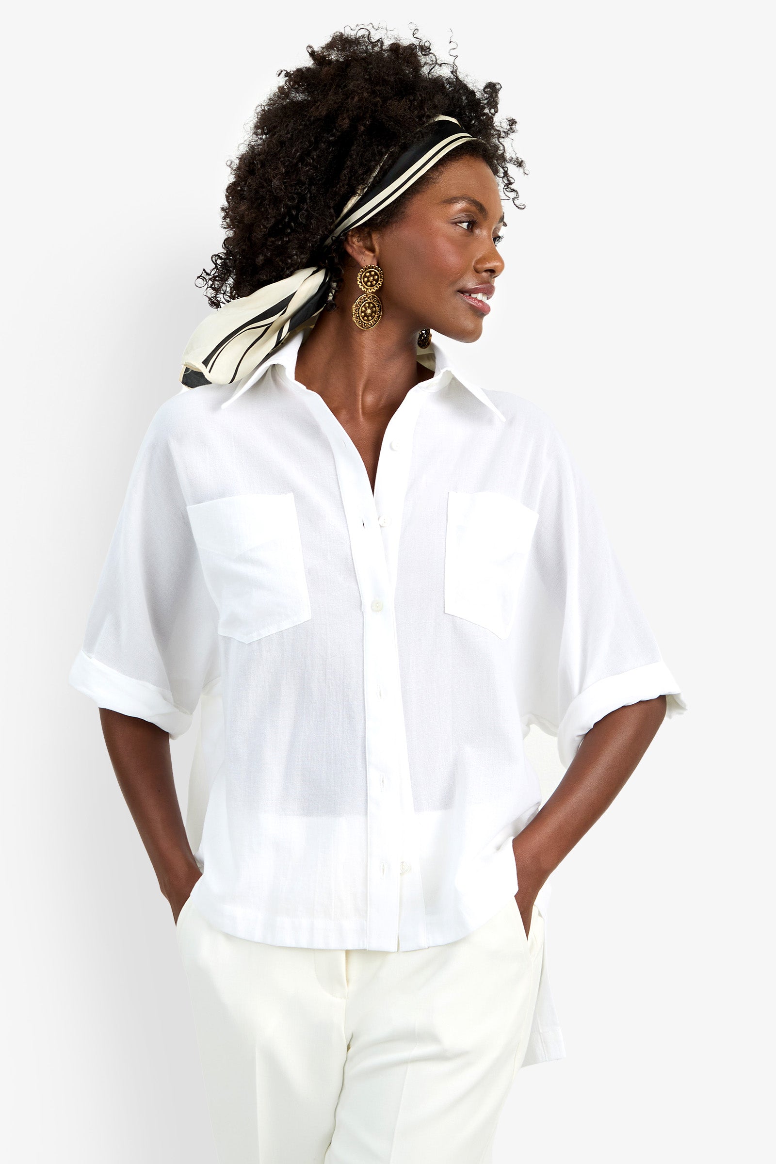 A woman wears the Hawaii Top—a loose white short-sleeve button-up with front pockets—paired with white pants. She has curly hair styled with a black-and-white scarf and accessorizes with large round earrings.