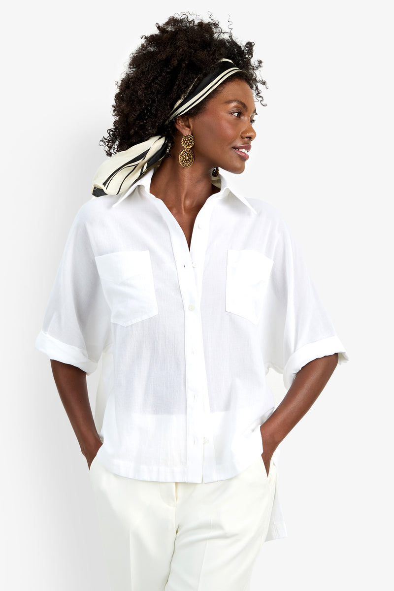 A woman wears the Hawaii Top—a loose white short-sleeve button-up with front pockets—paired with white pants. She has curly hair styled with a black-and-white scarf and accessorizes with large round earrings.