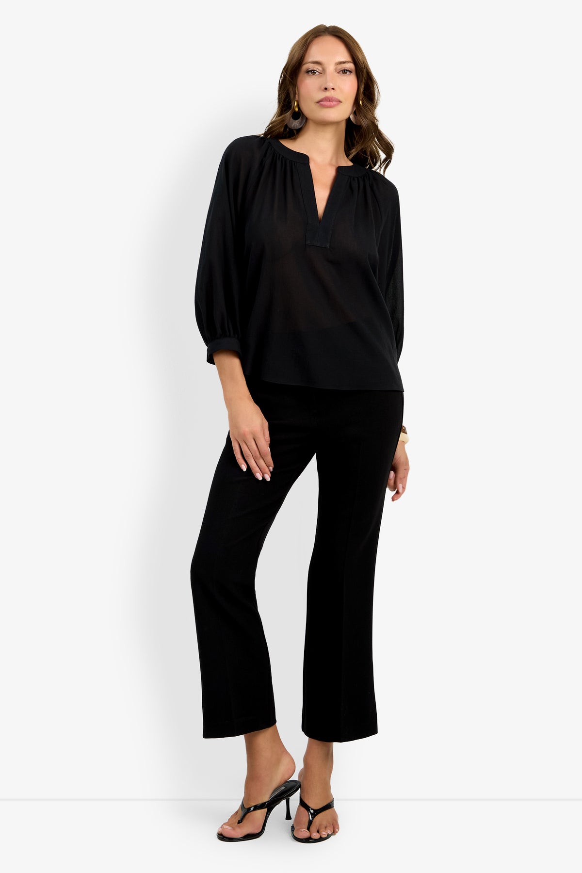 A woman with wavy brown hair and hoop earrings poses against a white background wearing the Light Hearted Top—a loose black blouse with three-quarter sleeves—paired with black pants and heeled sandals.