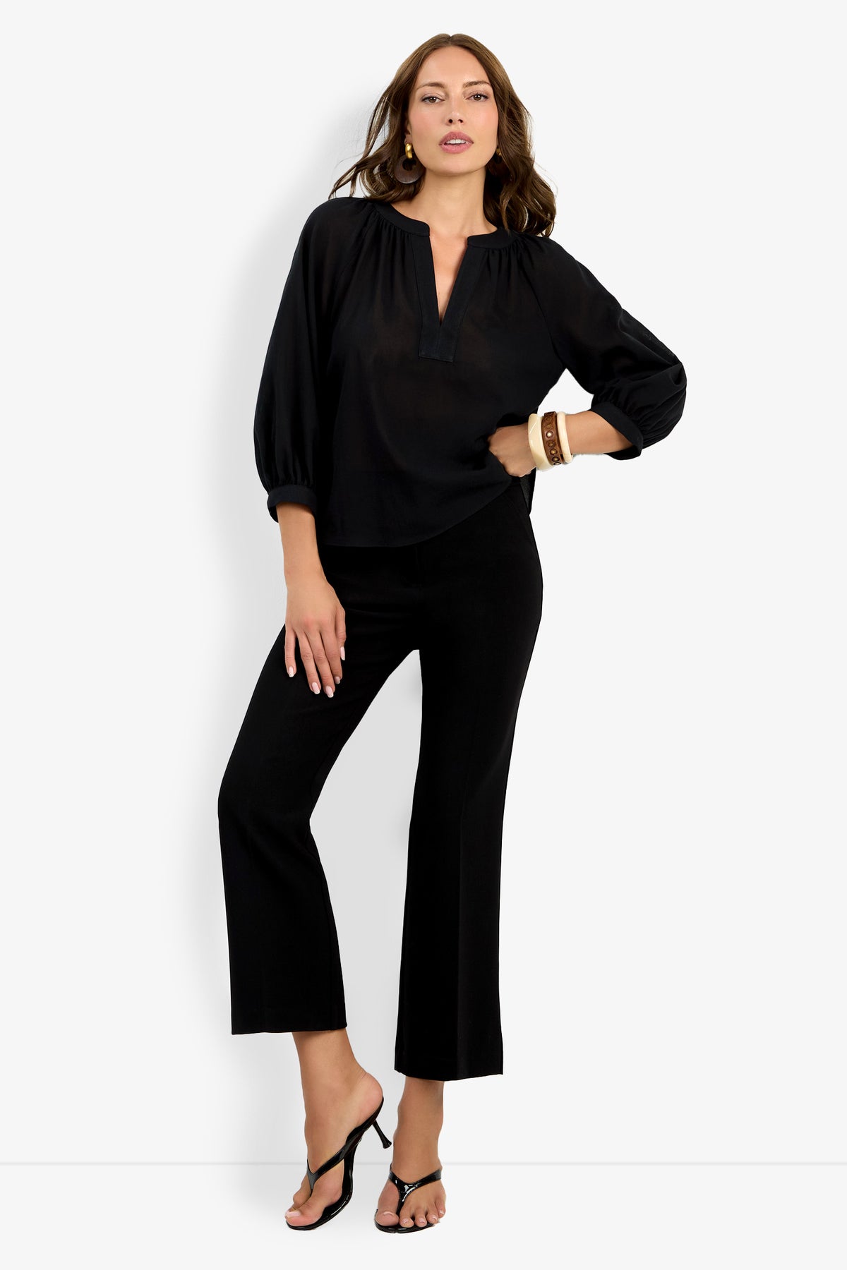 A woman stands against a white background wearing the Light Hearted Top, a loose black blouse with three-quarter sleeves, paired with black cropped pants, heeled sandals, and a brown bracelet, one hand on her hip and the other on her thigh.
