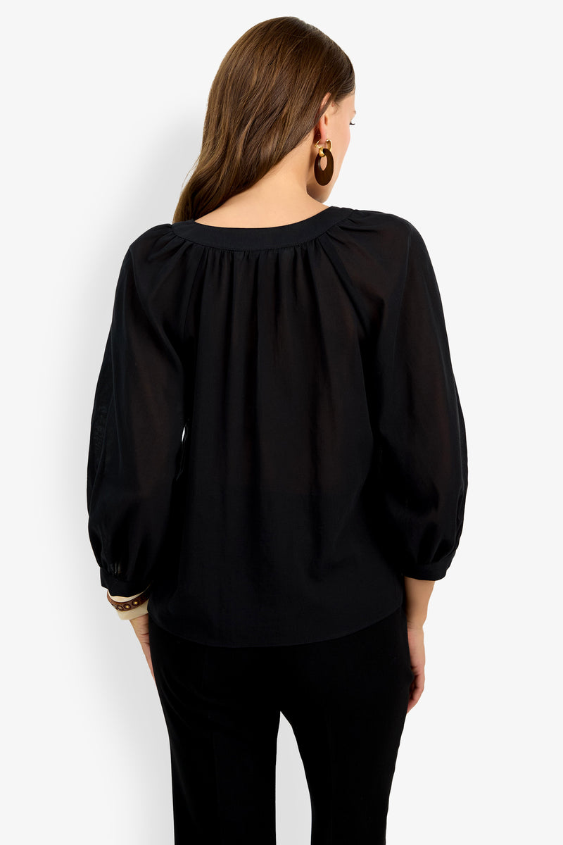 A woman with long brown hair is seen from the back wearing the Light Hearted Top, a loose black blouse with gathered details and balloon sleeves, paired with black pants. She accessorizes with hoop earrings and bracelets.