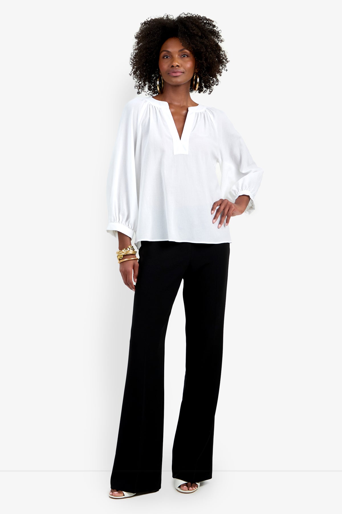 A woman stands confidently wearing the Light Hearted Top, a loose white V-neck blouse with long sleeves, paired with black wide-leg trousers, open-toed heels, gold bracelets, and earrings.