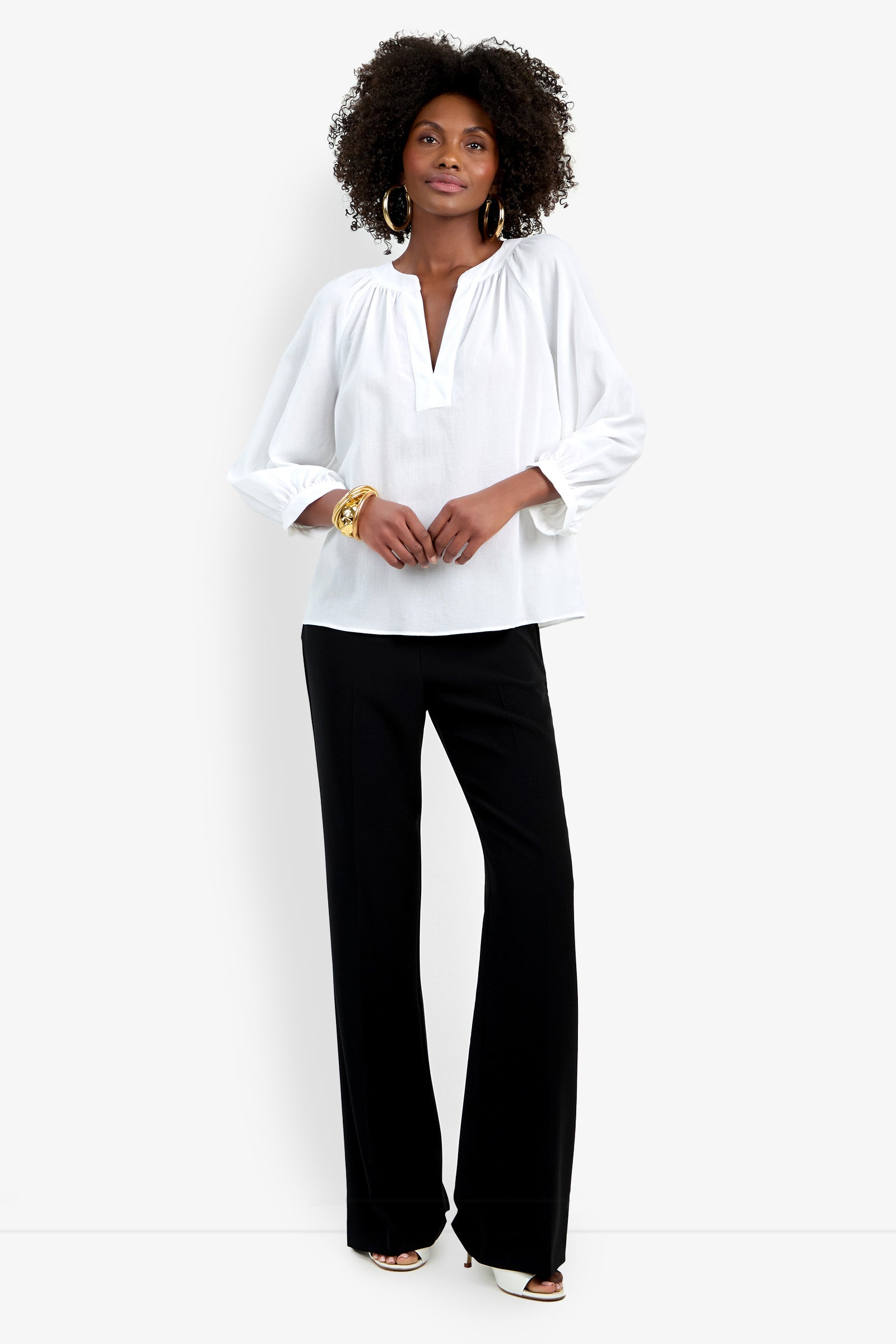 A woman stands against a white background wearing the Light Hearted Top, a loose white long-sleeve V-neck blouse, paired with black wide-leg pants. She completes the look with hoop earrings, bracelets, and white shoes.