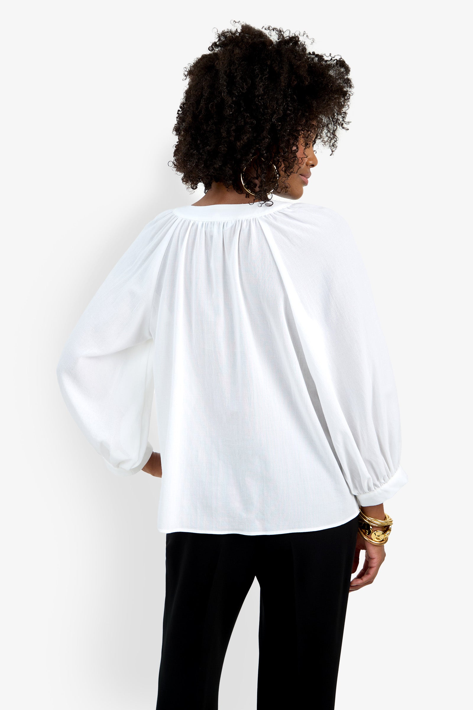 A woman with curly hair is seen from the back wearing the Light Hearted Top, a loose, long-sleeved white blouse, and black pants. She accessorizes with gold bracelets on her right wrist and stands against a plain white background.