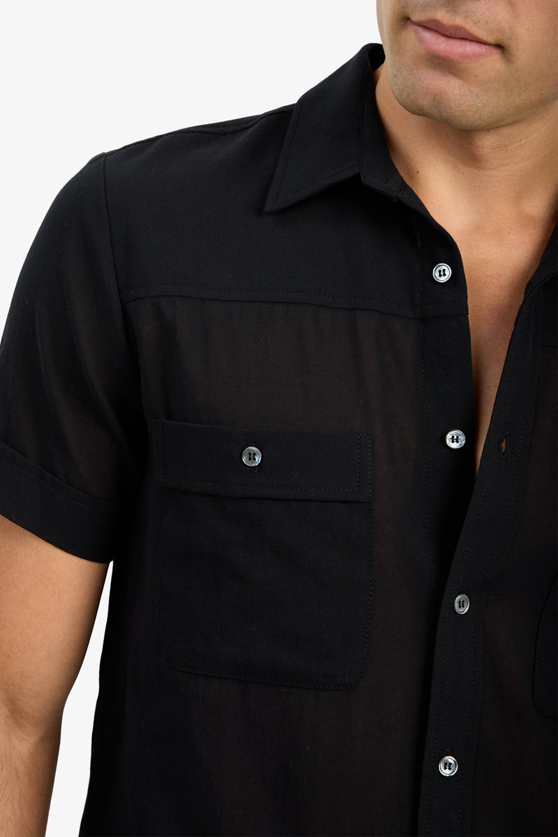 A person wearing the Will Shirt, a sheer black short-sleeve button-up with a front pocket, shown from the chest up against a plain background.