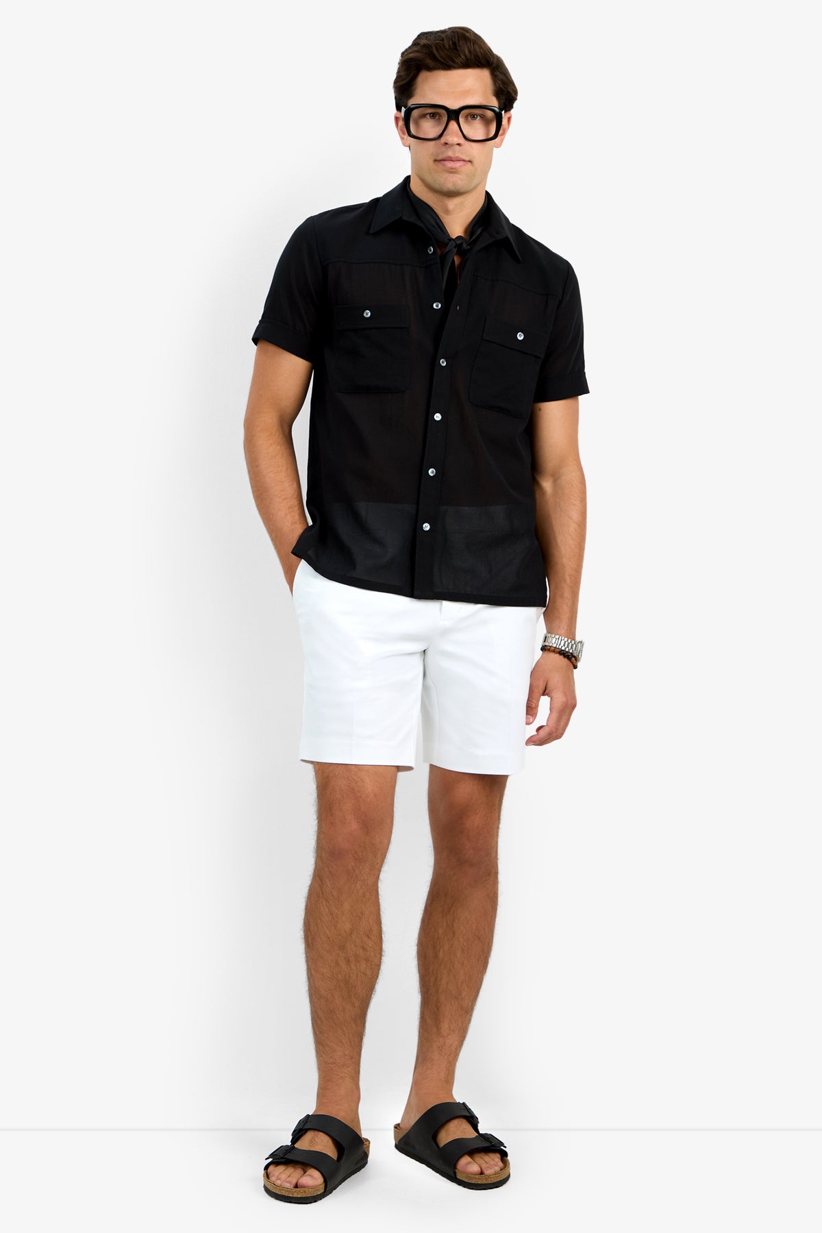 A man with a neutral expression stands against a white background wearing the black Will Shirt, white shorts, black sandals, and large black glasses, with one hand in his pocket.