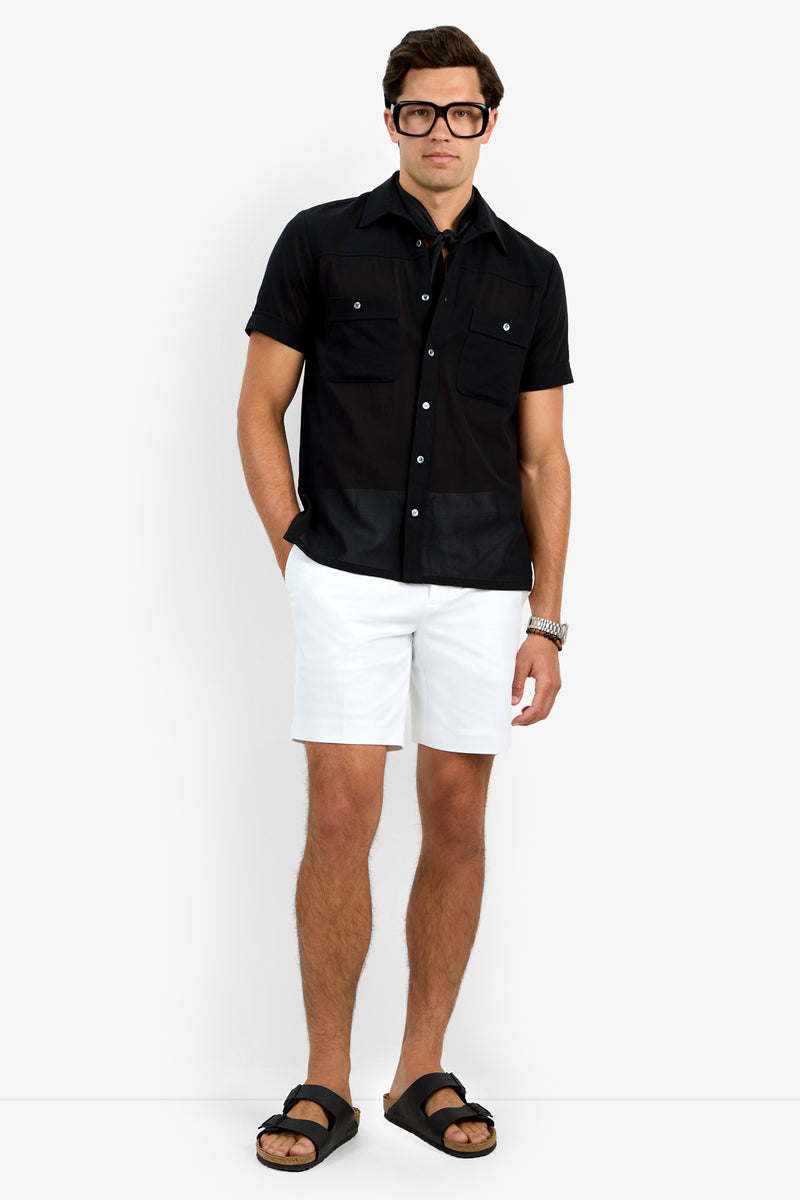 A man with a neutral expression stands against a white background wearing the black Will Shirt, white shorts, black sandals, and large black glasses, with one hand in his pocket.