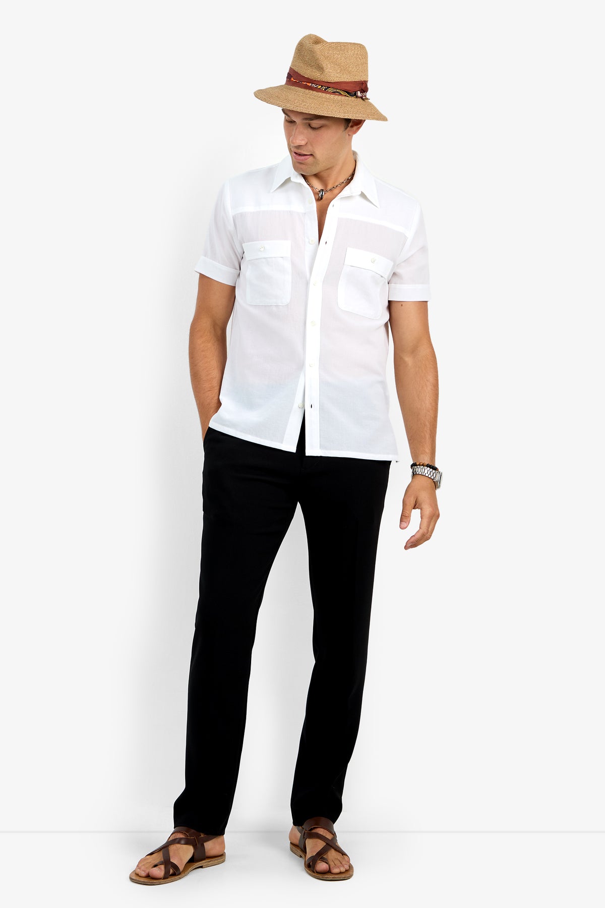 A man in the Will Shirt—short-sleeve, white, button-up—paired with black pants, brown sandals, and a straw hat stands against a white background with hands in pockets, looking down slightly.