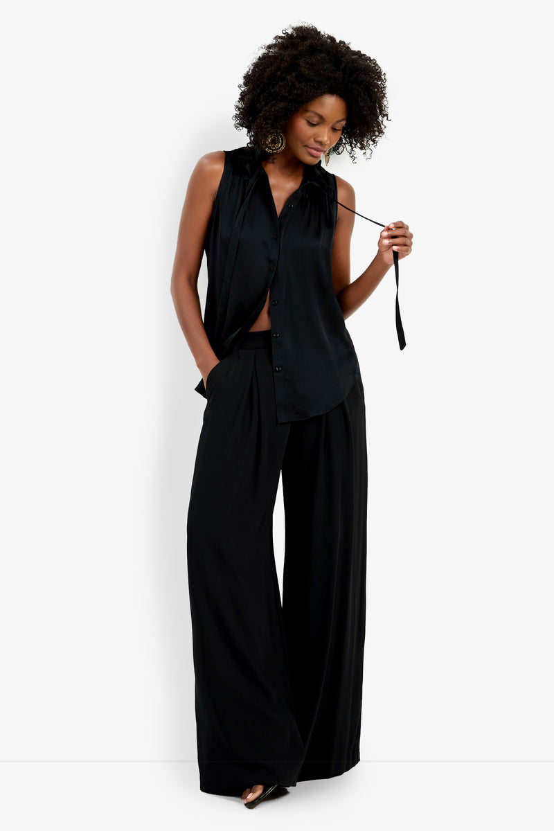 A woman wears the Cinzia Top, a sleeveless black button-up blouse, paired with wide-leg black pants. She stands with one hand in her pocket, holding her collar tie, and smiles slightly.