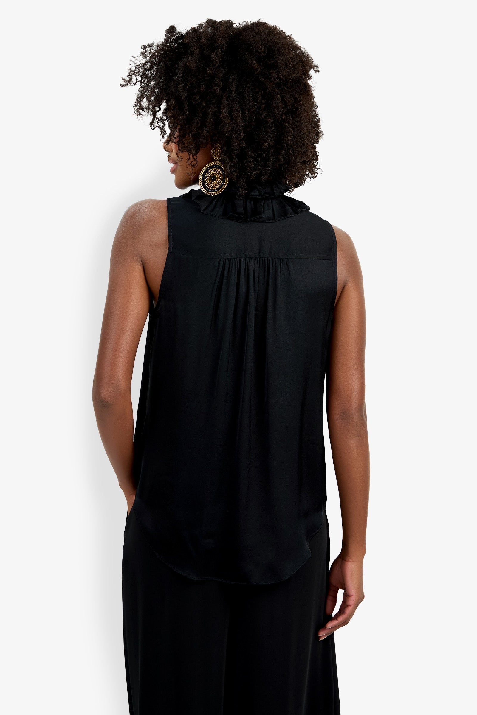 A woman with curly hair, wearing gold statement earrings and the sleeveless black Cinzia Top, stands with her back to the camera against a white background.