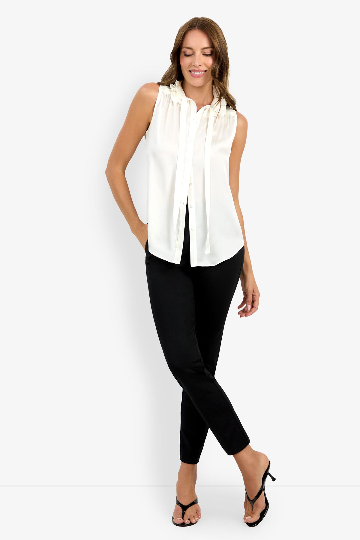 A woman stands against a white background, smiling with one hand in her pocket, wearing the Cinzia Top—a sleeveless white blouse with a tie-neck detail—paired with black slim-fit pants and black heeled sandals.