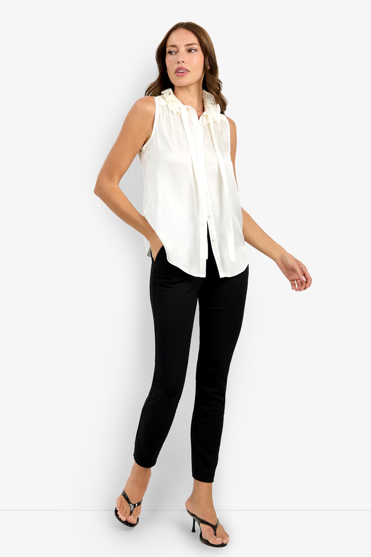 A woman models the Cinzia Top, a sleeveless white blouse with ruffle details, paired with black slim pants and black heeled sandals, standing against a plain white background with one hand in her pocket.