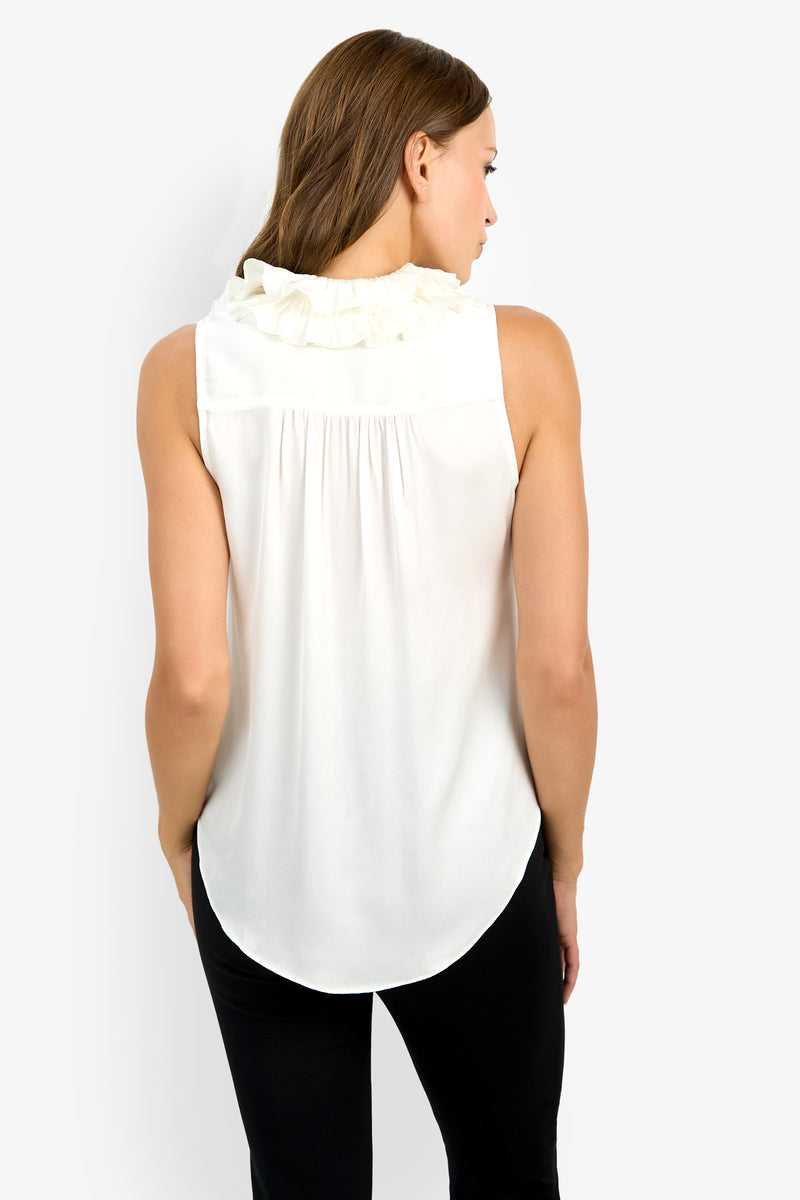 A woman with brown hair is seen from behind wearing the Cinzia Top, a sleeveless white blouse featuring ruffle details on the collar, paired with black pants, standing against a plain white background.