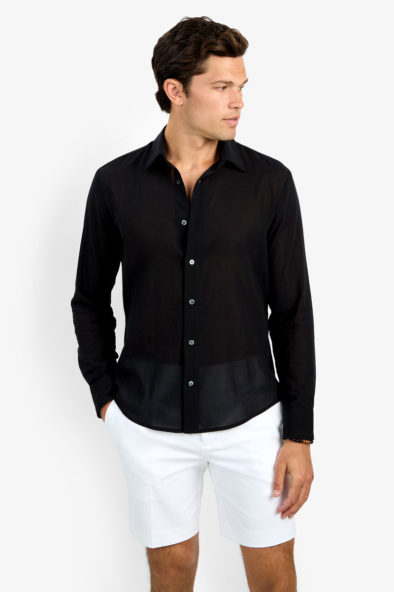 A man stands against a white background wearing the black Aaron Shirt with long sleeves and white shorts. He has one hand in his pocket and is looking to his left.