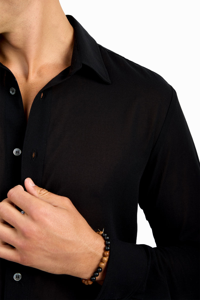 A person wearing the Aaron Shirt, a black button-up, holds it near their chest with their left hand. They have a brown and black beaded bracelet on their wrist. The background is plain white.