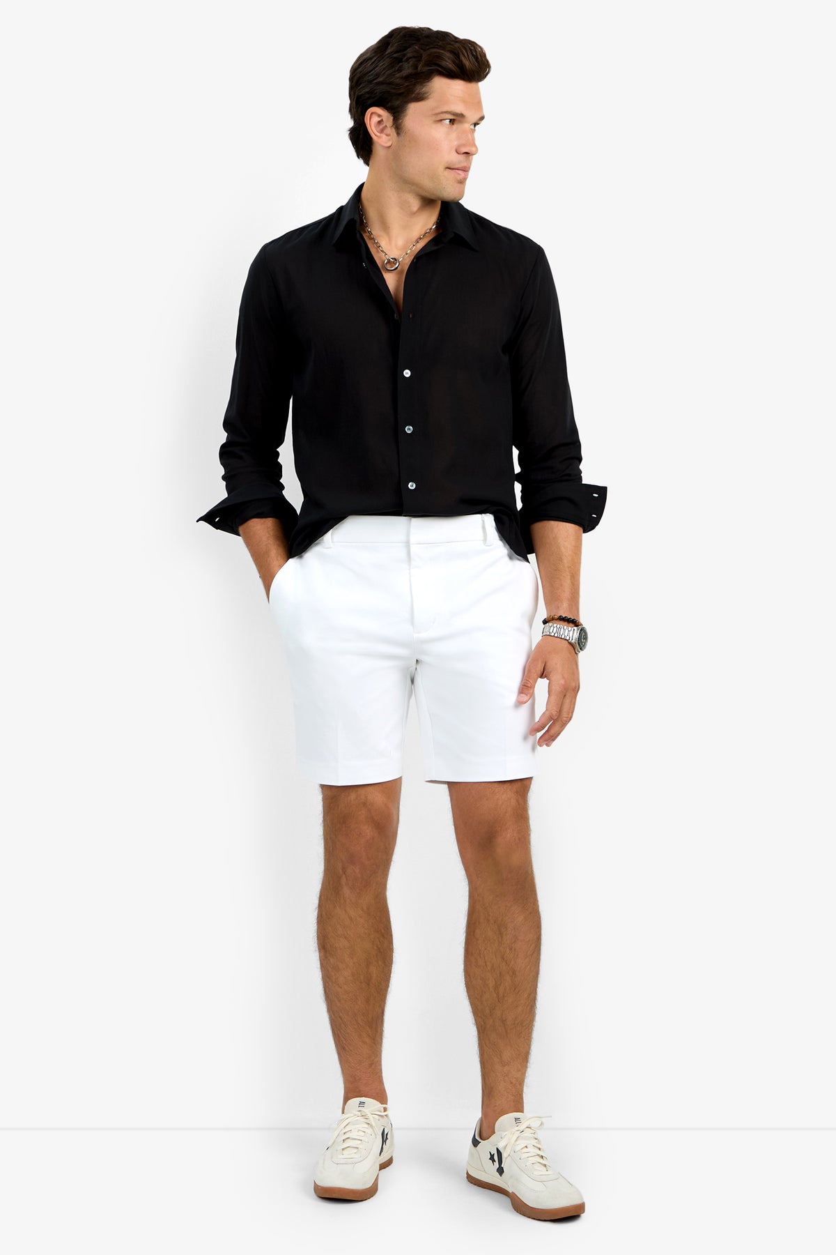 A man stands against a plain white background wearing the black Aaron Shirt, white shorts, two layered necklaces, a wristwatch, and white sneakers with black details. His hands are in his pockets as he looks to his left.