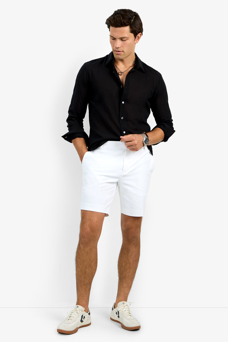 A man stands with hands in pockets, wearing the black Aaron Shirt, white shorts, white sneakers with black accents, a watch, and layered necklaces against a plain white background.