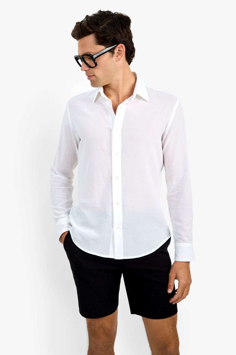 A man in the Aaron Shirt, a white button-up, and black shorts looks to the side with one hand in his pocket. He has short dark hair, large black glasses, and stands against a plain white background.