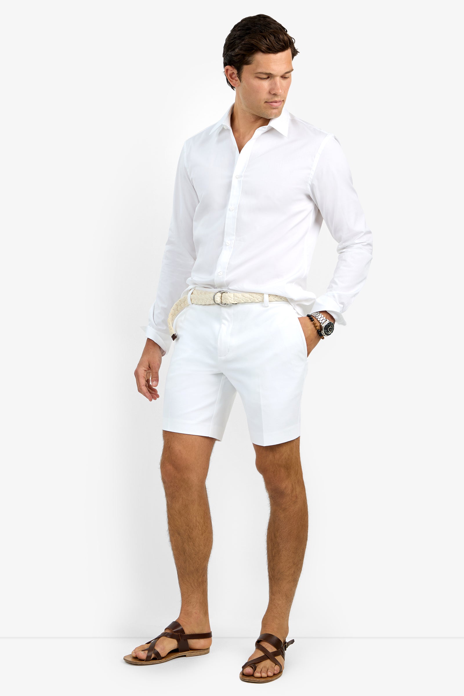 A man with short dark hair stands looking left, wearing the white Aaron Shirt, white shorts, a braided belt, a watch, and brown sandals. One hand is in his pocket.