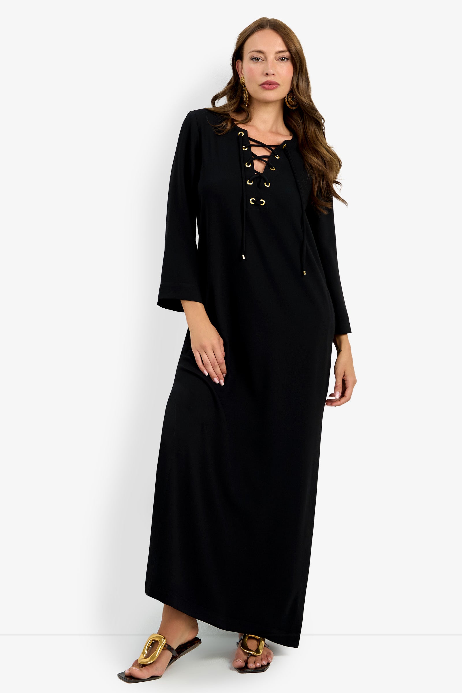 A woman wearing the Catamaran Dress, a long black dress with gold grommet lace-up detail at the neckline and long sleeves, paired with gold sandals, stands against a plain white background.
