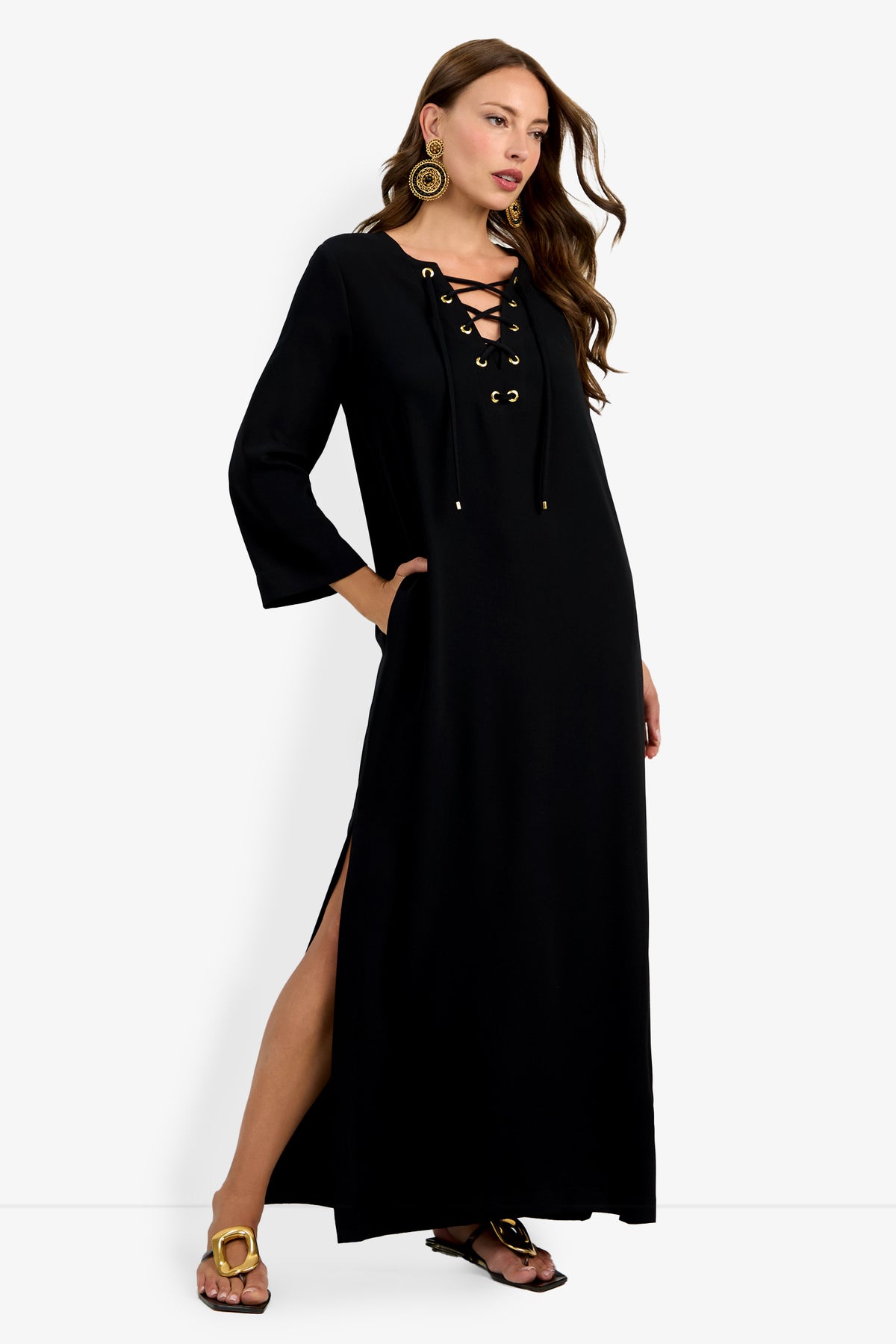 Woman wearing the Catamaran Dress, a long black dress featuring lace-up front detail, three-quarter sleeves, side pockets, and a slit. She accessorizes with large round earrings and black sandals with gold accents.