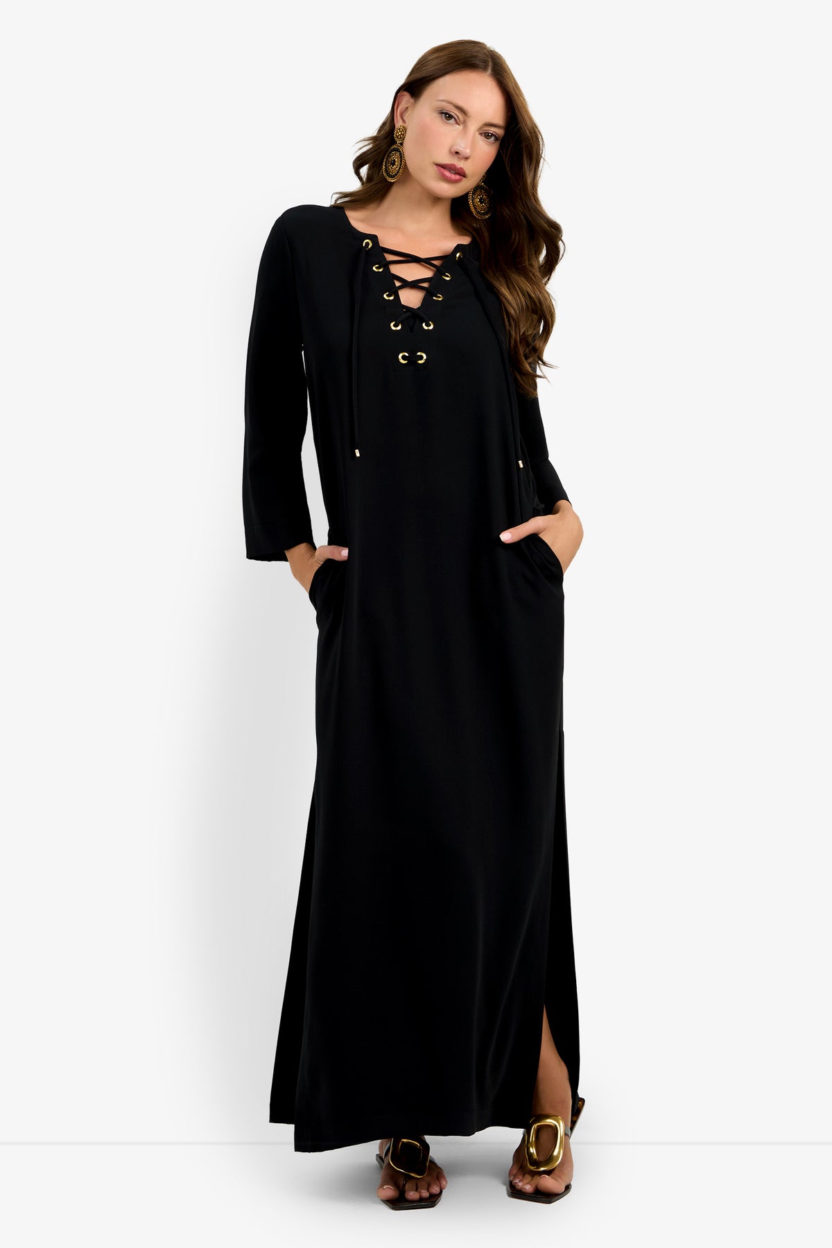 A woman wears the Catamaran Dress—a long black dress with a lace-up neckline, three-quarter sleeves, side pockets, and a hem slit. She stands with her hands in her pockets, accessorized with hoop earrings and sandals.